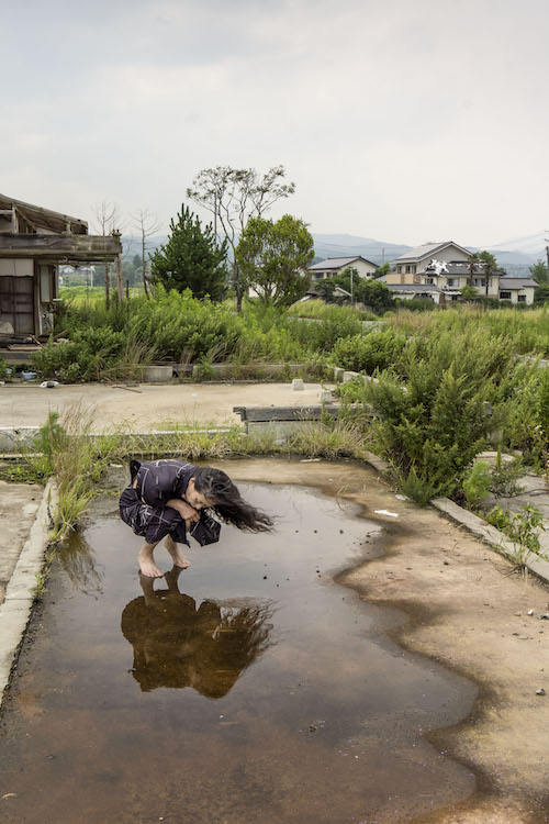 Eiko crouches in a puddle on a concrete square in a barren Fukushima